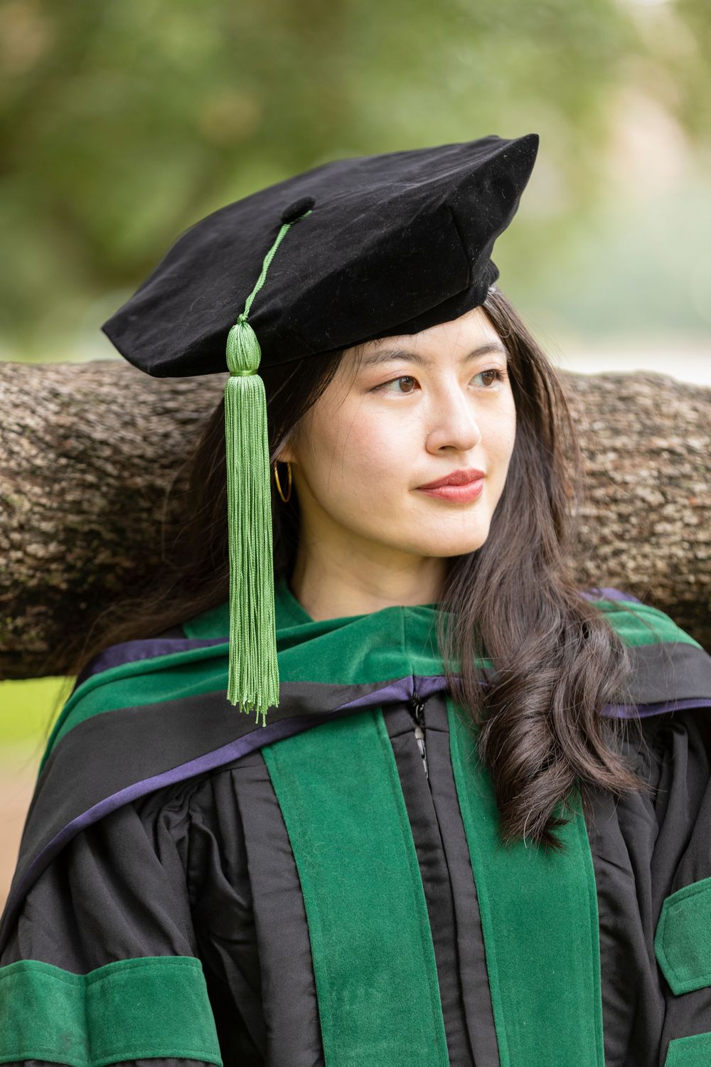 Young woman in black mortarboard and green doctoral graduation gown leaning against a tree outdoors.