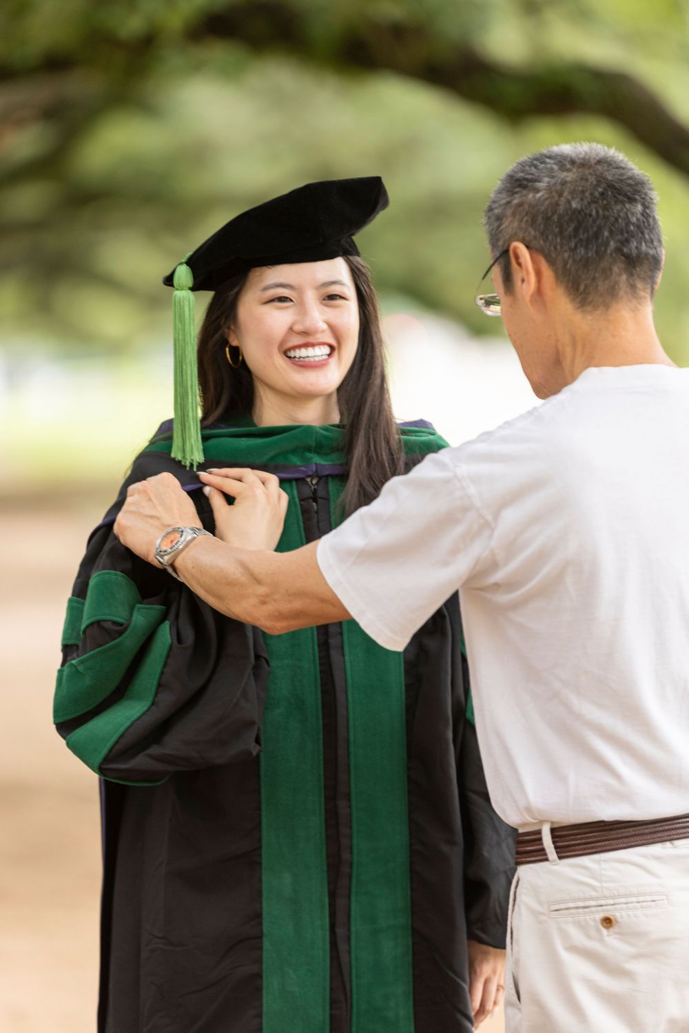 Smiling woman in black and green graduation gown and cap receives help adjusting her robe outdoors.