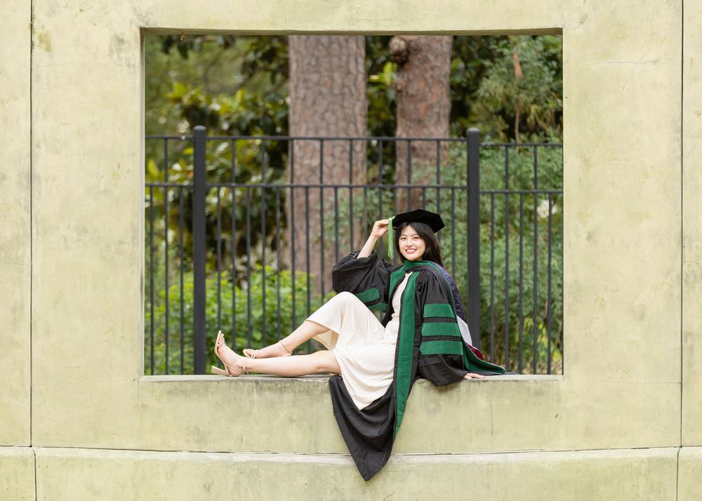 Woman in black graduation gown and cap sits on stone wall ledge, smiling outdoors near iron fence.