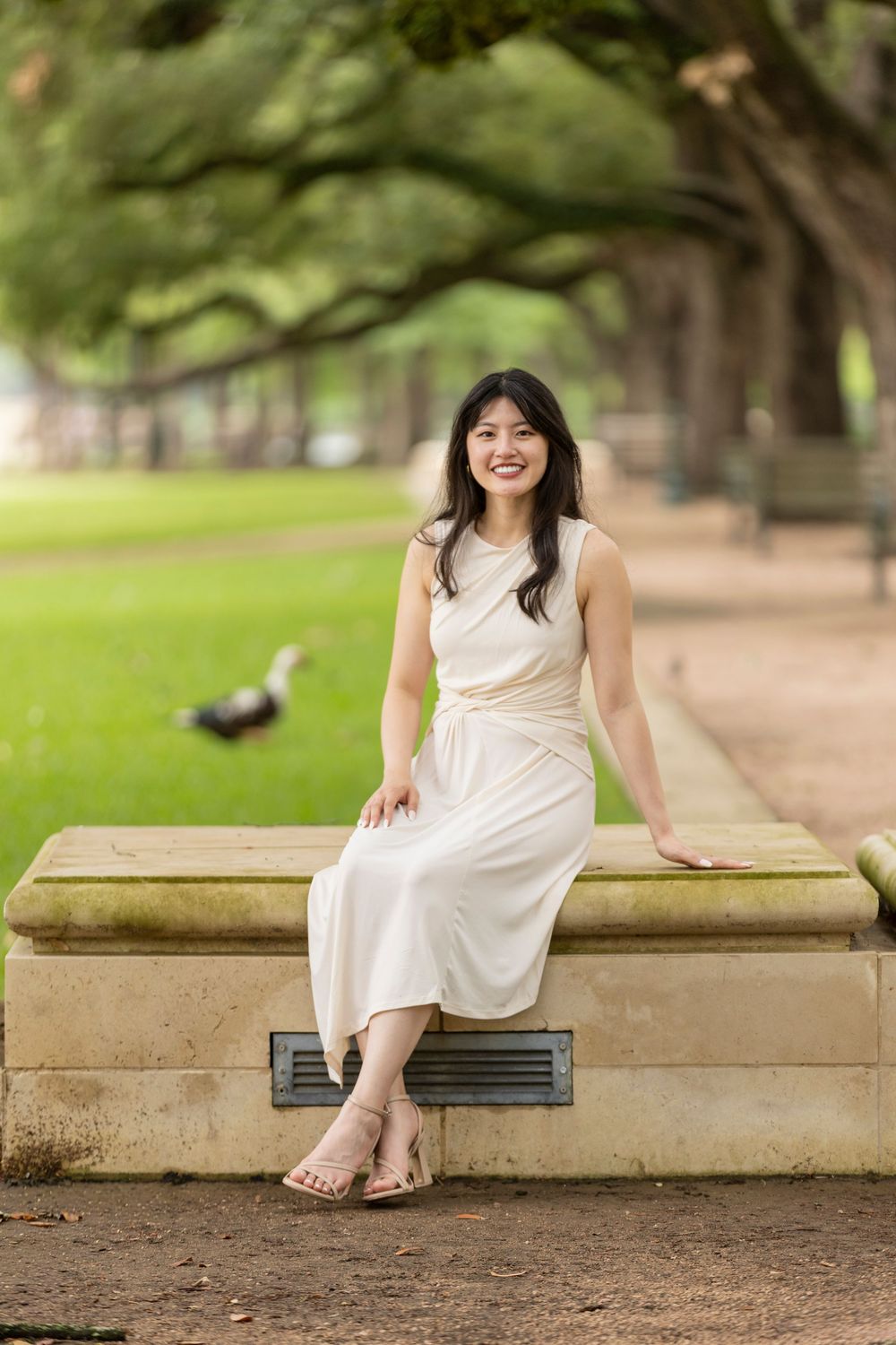 Smiling young woman in white dress sitting on stone bench in lush green park with trees.