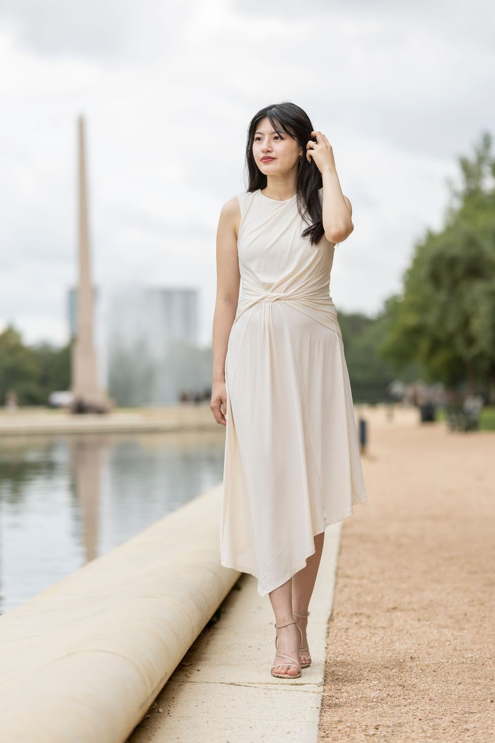 Young woman in elegant cream asymmetric dress poses by a fountain in a park on a cloudy day.