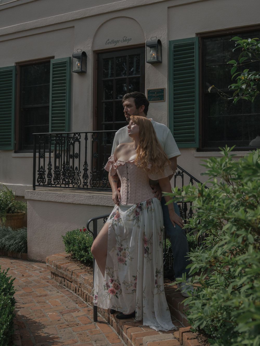 Couple standing on brick steps outside a charming cottage with green shutters and lush garden surroundings.