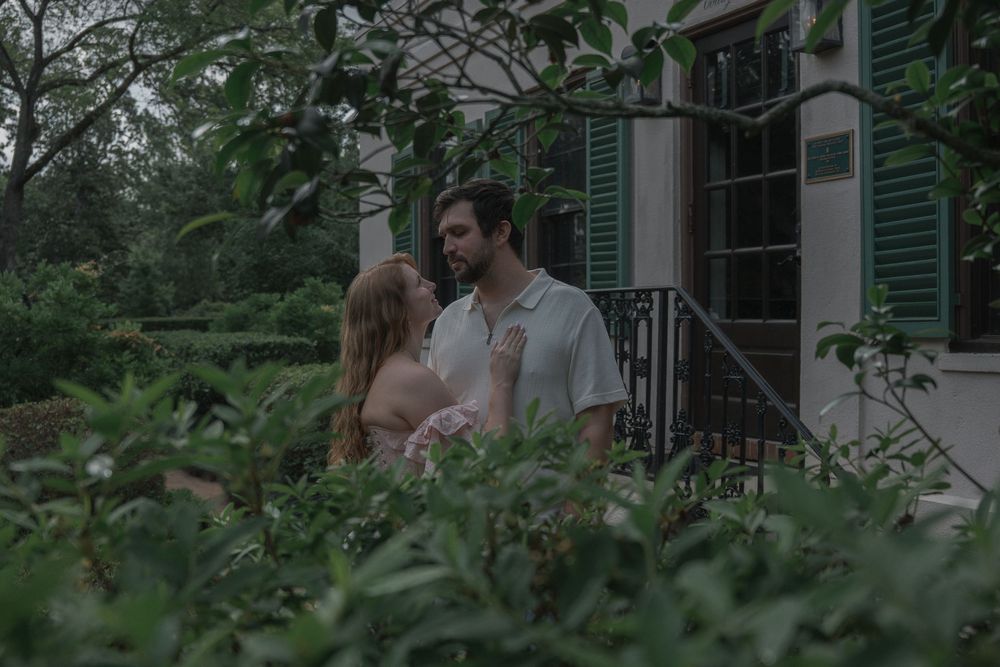 Couple sharing a romantic moment among lush greenery near a historic home with green shutters.
