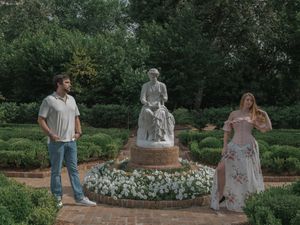 Couple standing beside an elegant garden statue surrounded by manicured hedges and white flowers.