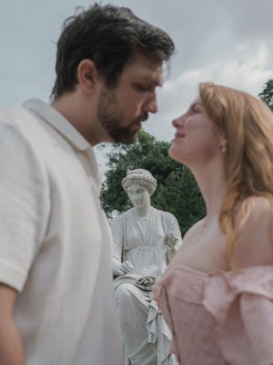 Couple gazing into each other's eyes with a classical stone statue between them in a garden.