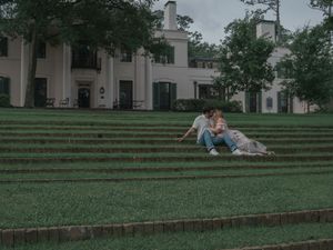Couple sitting together on terraced grass steps in front of a grand white estate home.
