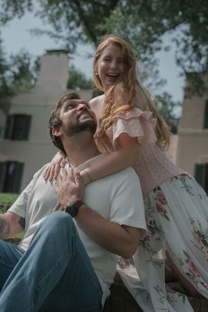 Joyful woman sitting behind man outdoors, laughing while hugging him beneath a shady tree.