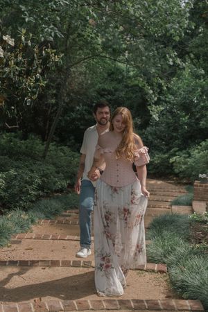 Couple walking hand in hand along a brick garden path surrounded by lush green trees.