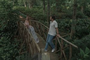 Couple posing on a wooden bridge in a lush forest, woman seated on the railing.