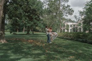 Man lifting woman in his arms on a green lawn with a large estate house visible behind them.