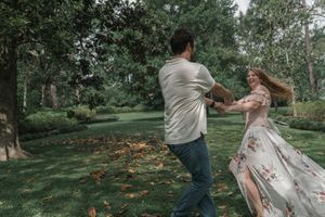 Couple dancing and spinning joyfully on a green lawn surrounded by tall trees in a garden.