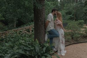 Couple sharing a kiss while leaning against a tree on a scenic garden path with wooden fence.