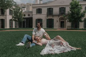 Couple sitting on lawn in front of elegant mansion, woman resting on man's lap.