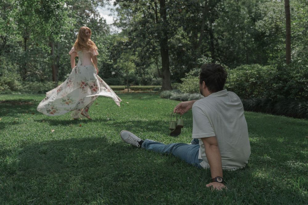Woman in floral dress twirling on grass while man sits watching in sunny park.
