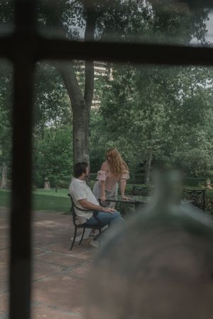 Man sitting on bench watching woman stand by stone wall in lush garden setting.