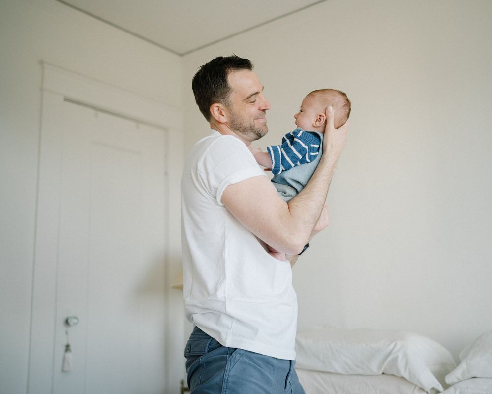 A father lovingly holds his newborn baby up in a bright white bedroom, smiling warmly at the infant.