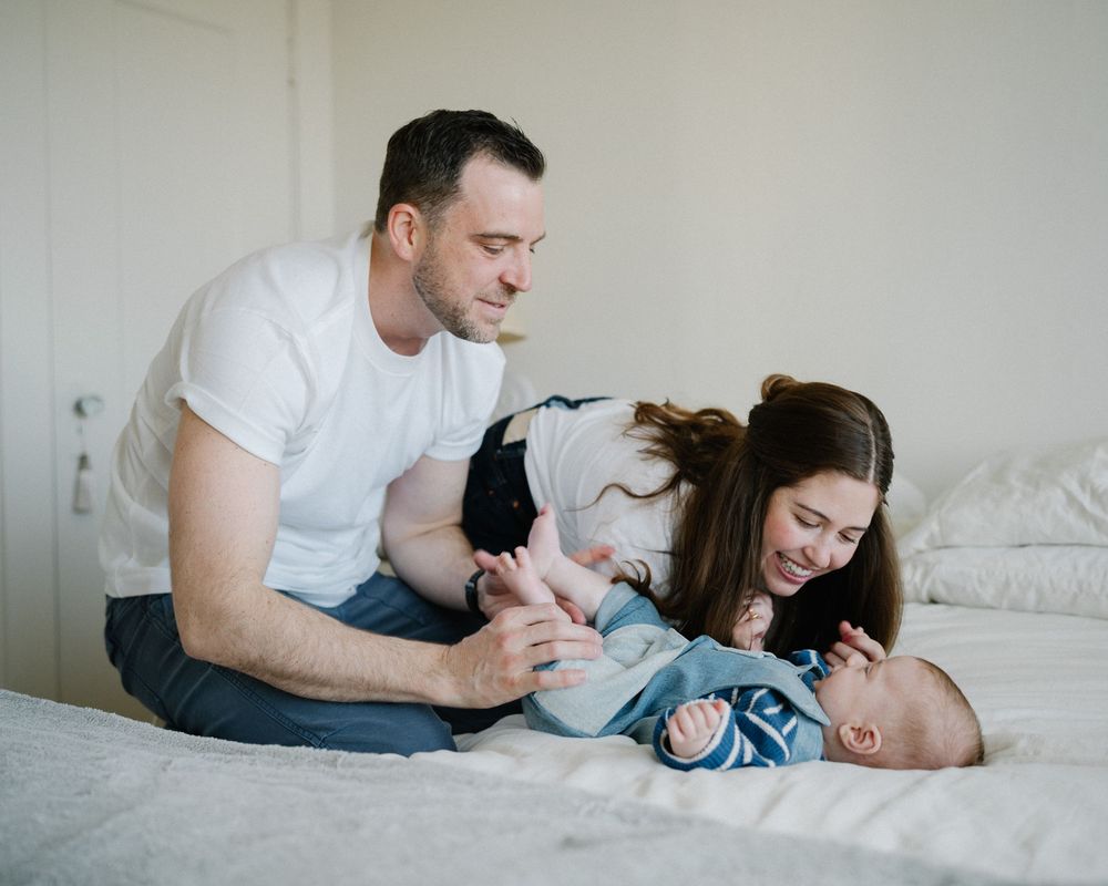 Parents smiling and playing with their newborn baby on a white bed in a bright bedroom.
