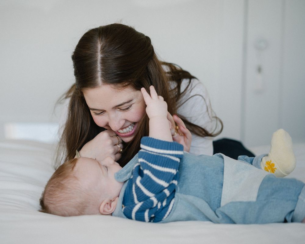 A smiling mother gazes lovingly at her baby boy lying on a white bed, sharing a tender moment.