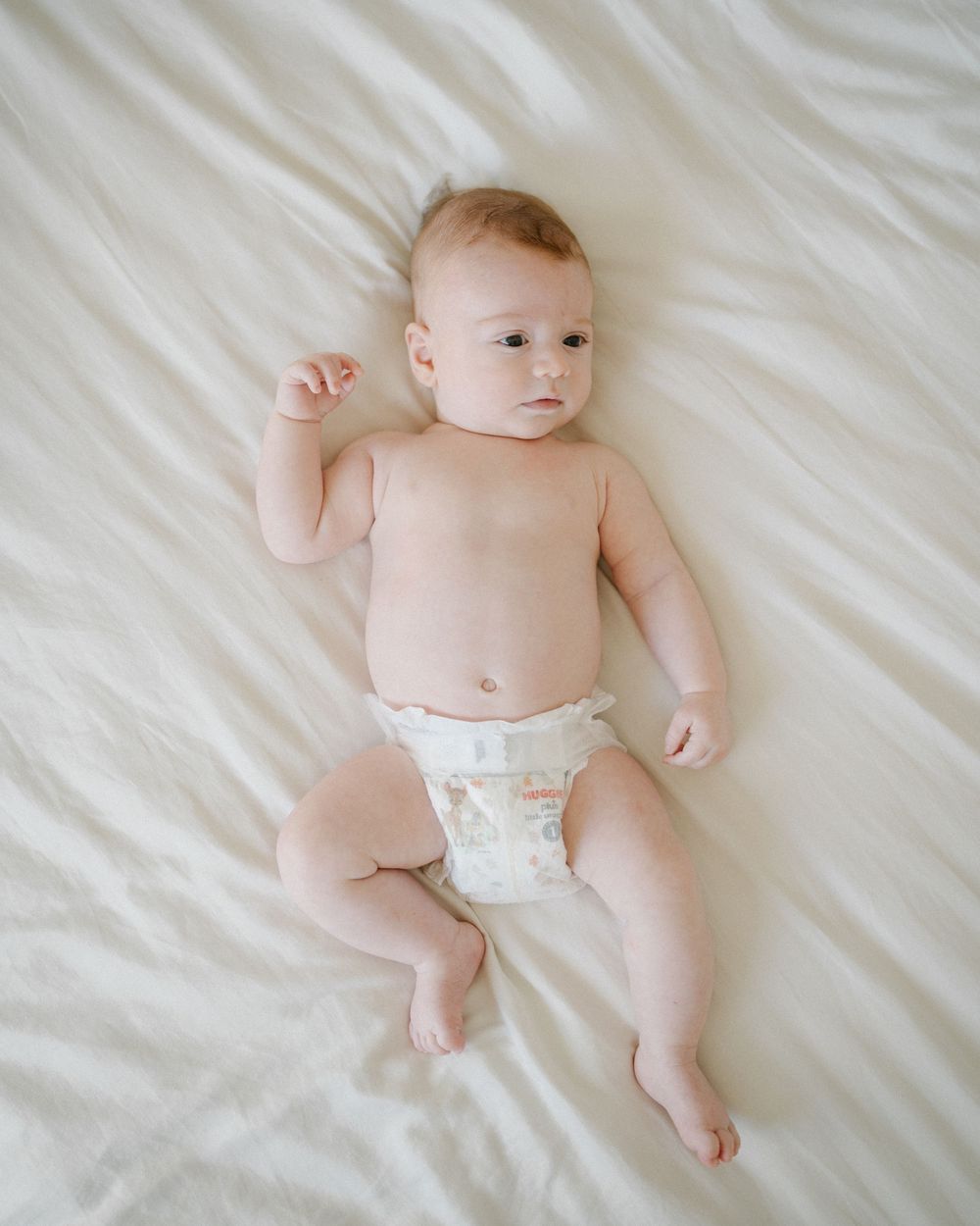 Adorable baby lying on white bedding wearing a diaper, looking up with one arm raised.