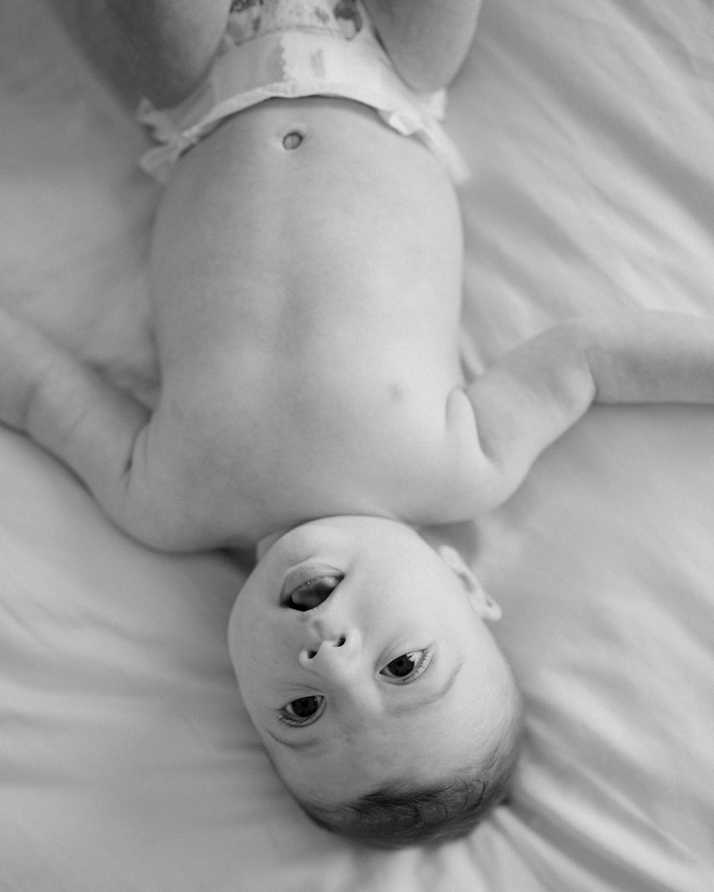 Black and white photo of a newborn baby lying on its back on white bedding, looking up at the camera.