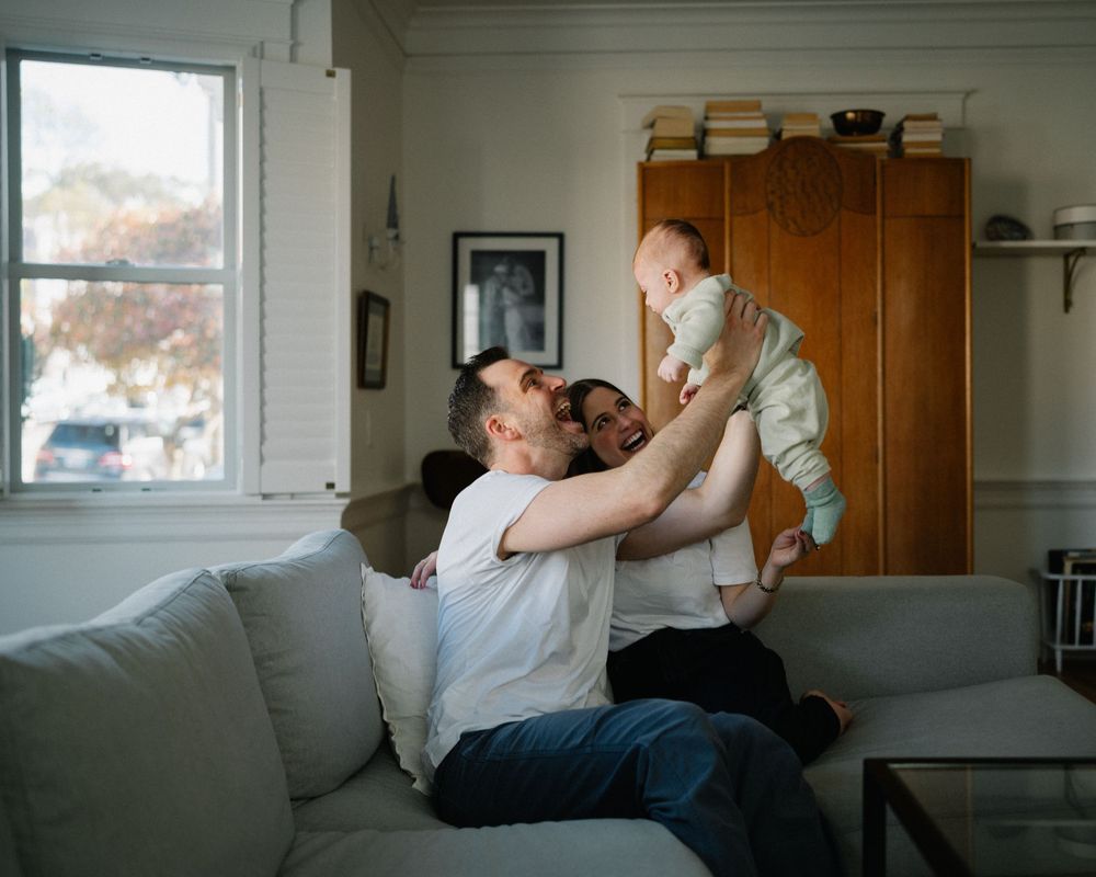 Happy couple lifting their baby in the air while sitting on a gray sofa in a cozy living room.