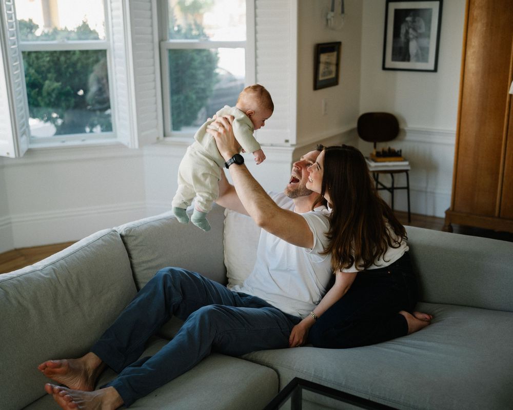 A mother lying on a gray sofa joyfully lifts her smiling baby in a bright, cozy living room.