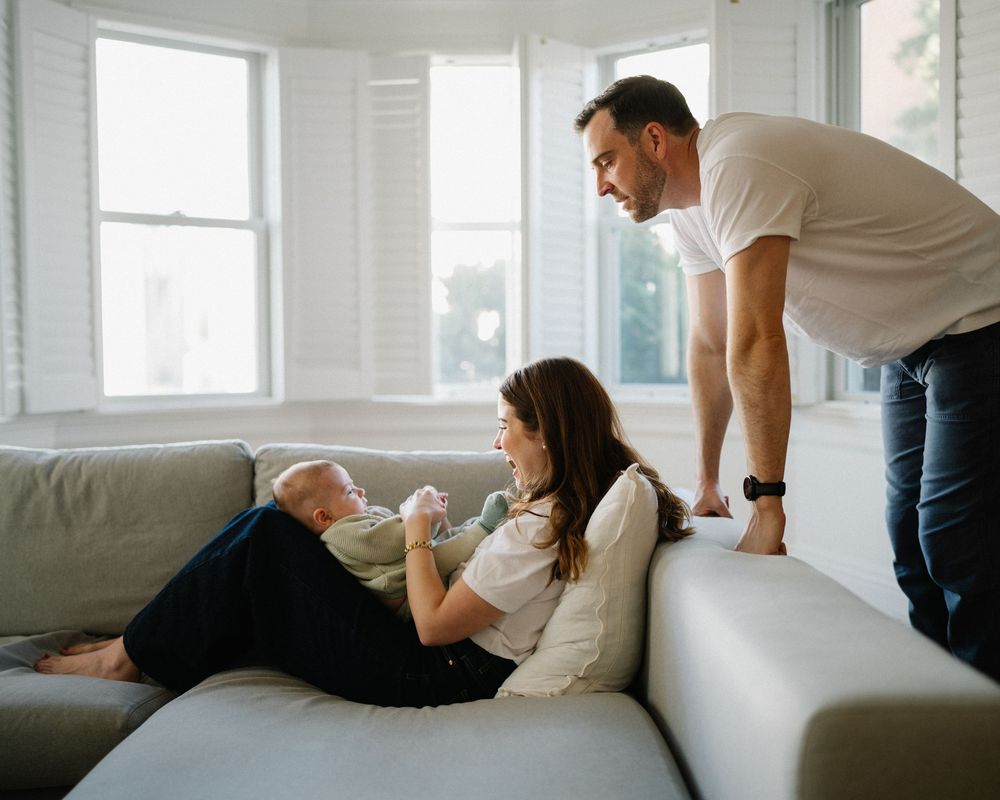 Mother reclining on sofa holding newborn while father leans over smiling in a bright living room.