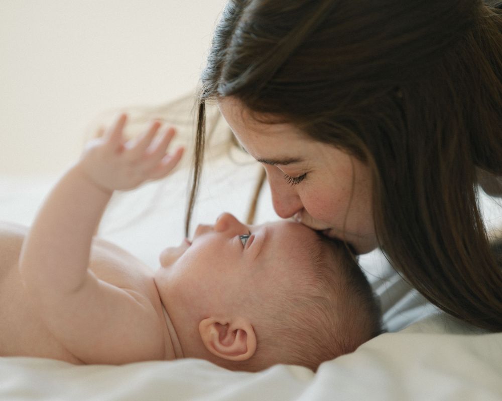 A loving mother gently kisses her newborn baby lying on a white bed in a tender, intimate moment.