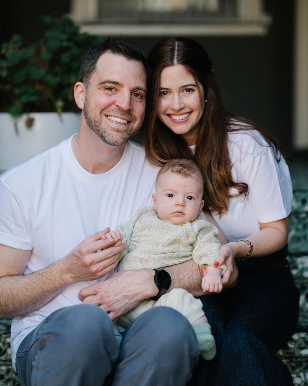 A smiling couple sits outdoors holding their infant baby dressed in a cream outfit, posing for a family portrait.