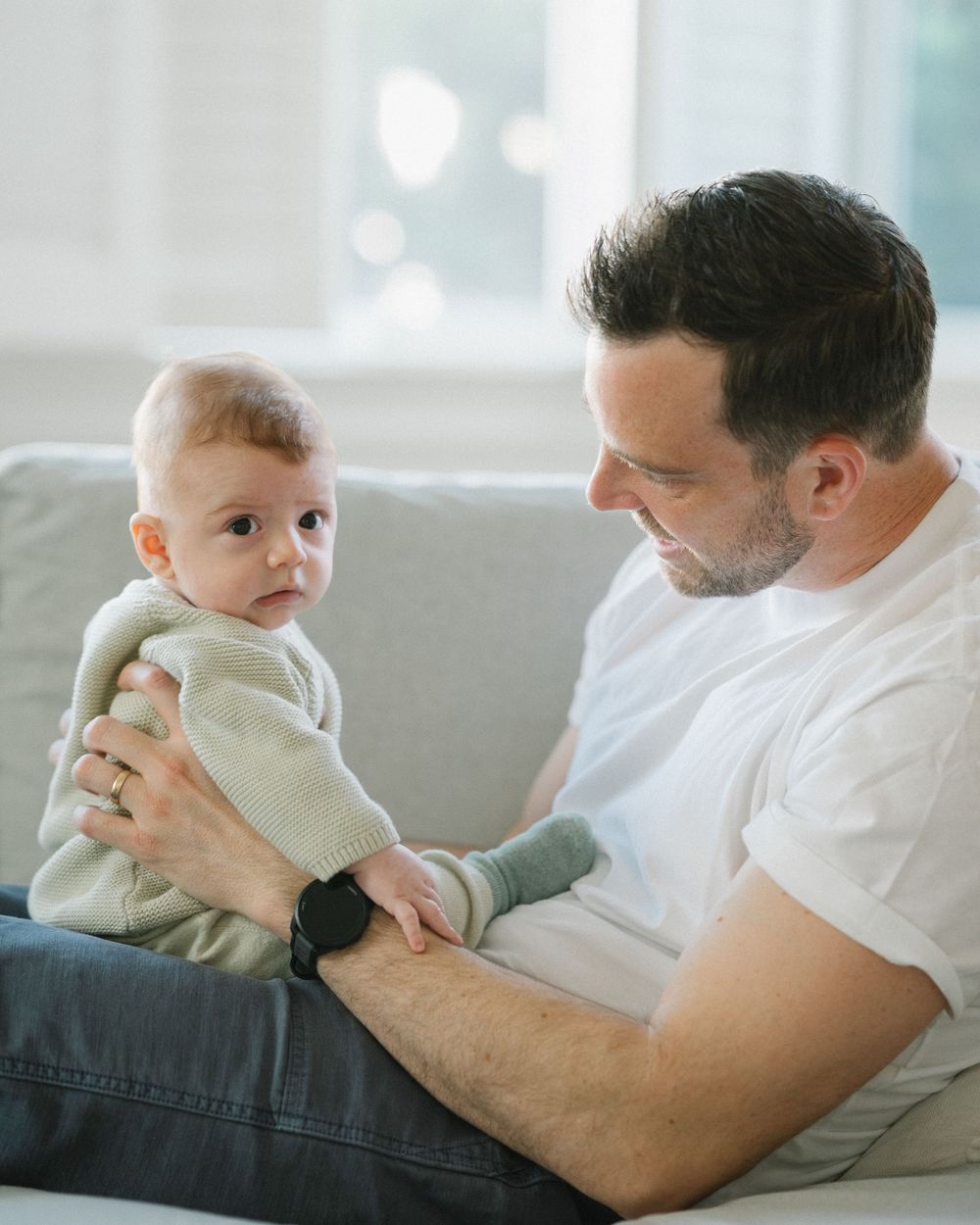 A father sits on a sofa holding his baby, gazing down lovingly while the infant looks at the camera.