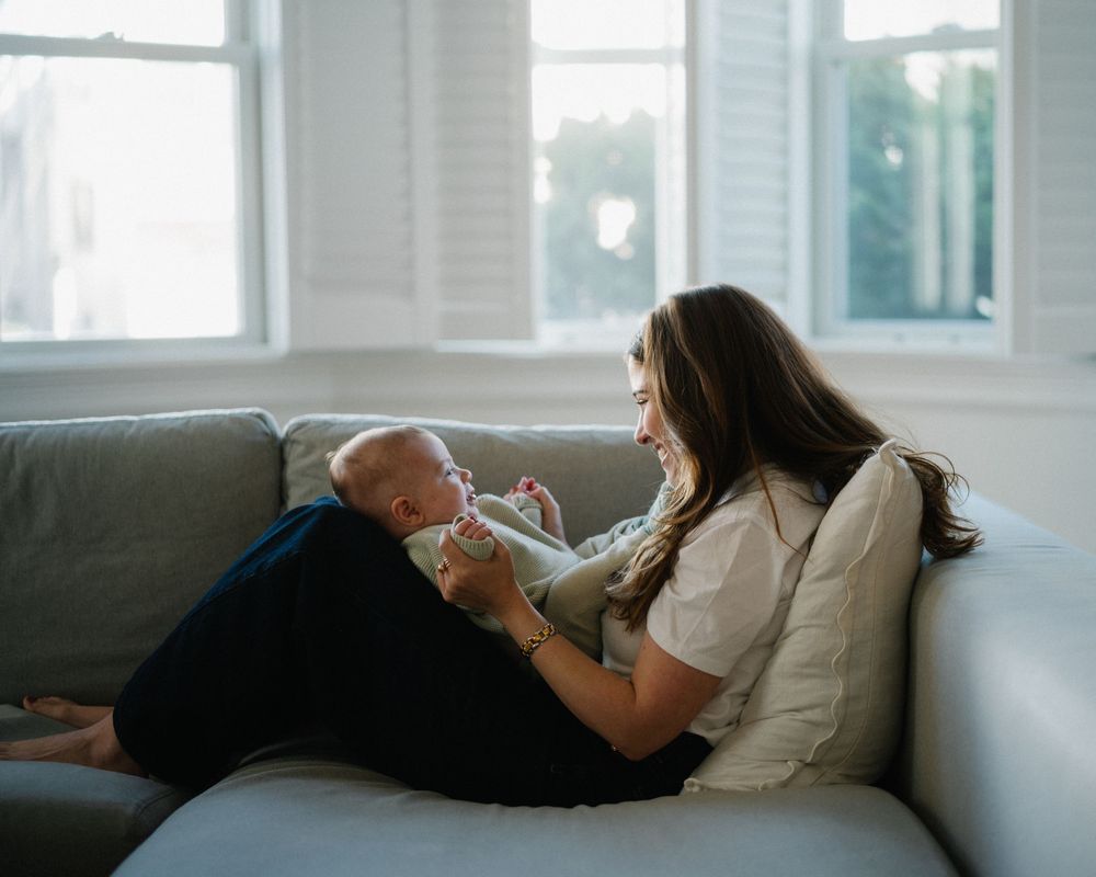 Young mother relaxing on gray sofa, lovingly interacting with her infant baby by bright windows.