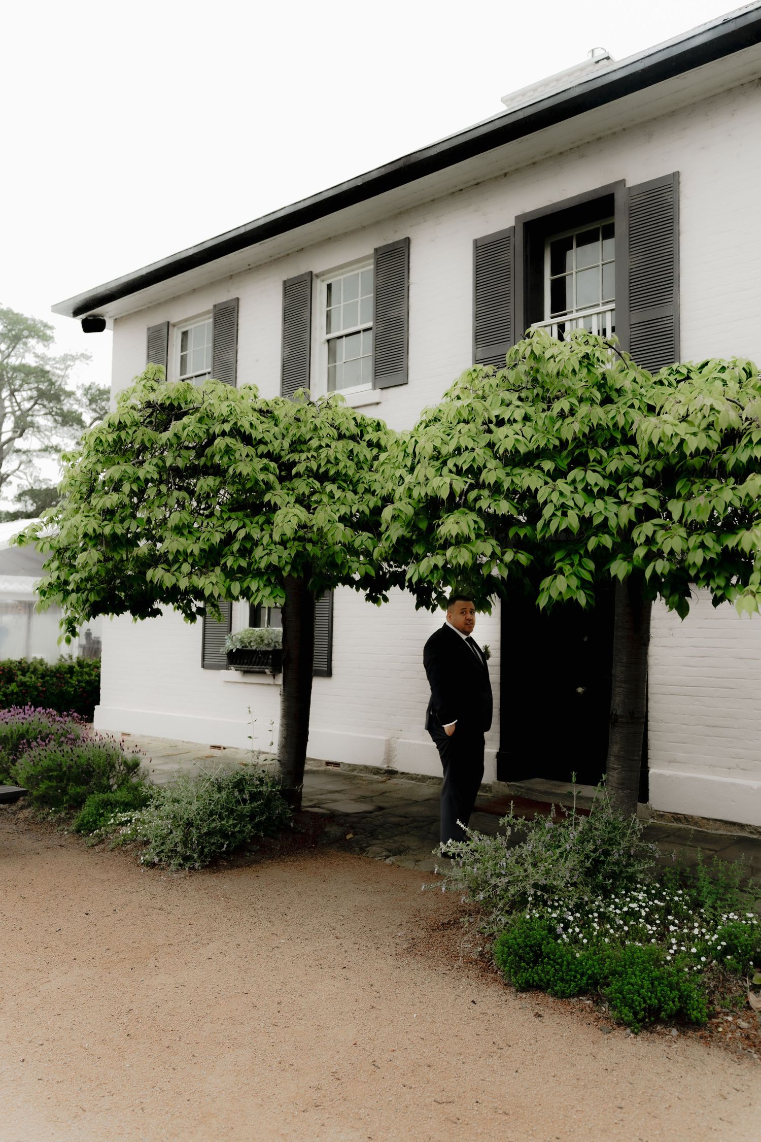 Person in formal attire stands beside espaliered trees against a white colonial building with gray shutters.