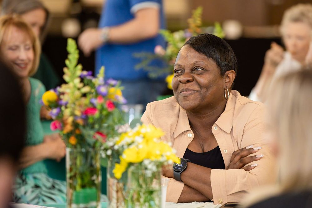Smiling Black woman in beige jacket sits at a table with colorful floral arrangements at a social gathering.