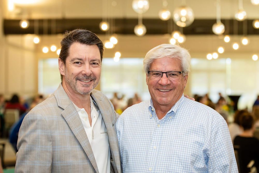 Two smiling men pose together at a bright indoor event with hanging lights and guests in the background.