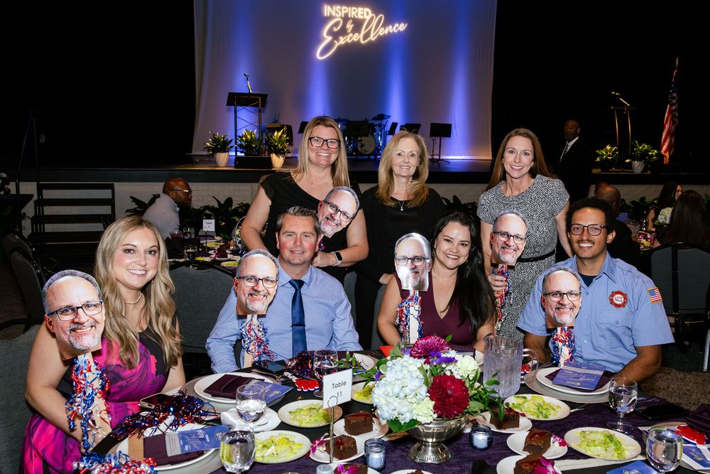 Group of smiling guests seated and standing at a gala dinner table with floral centerpieces and stage backdrop.