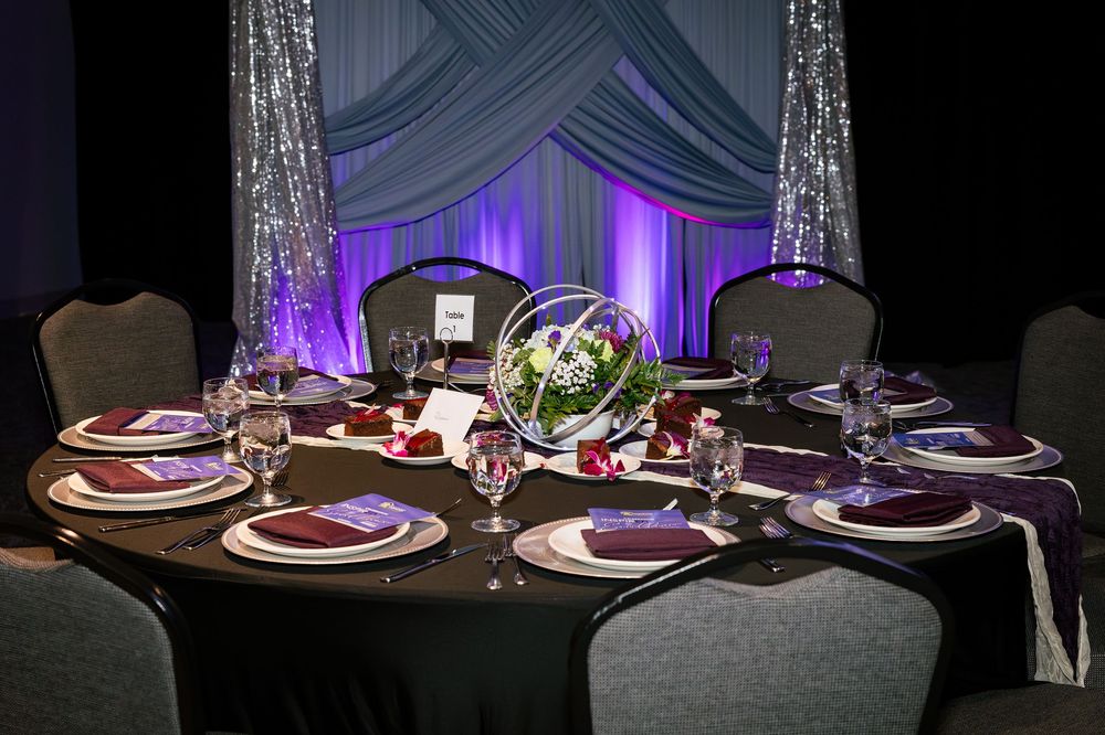 Elegant banquet table with purple napkins, floral centerpiece, and sparkling backdrop for a formal event.