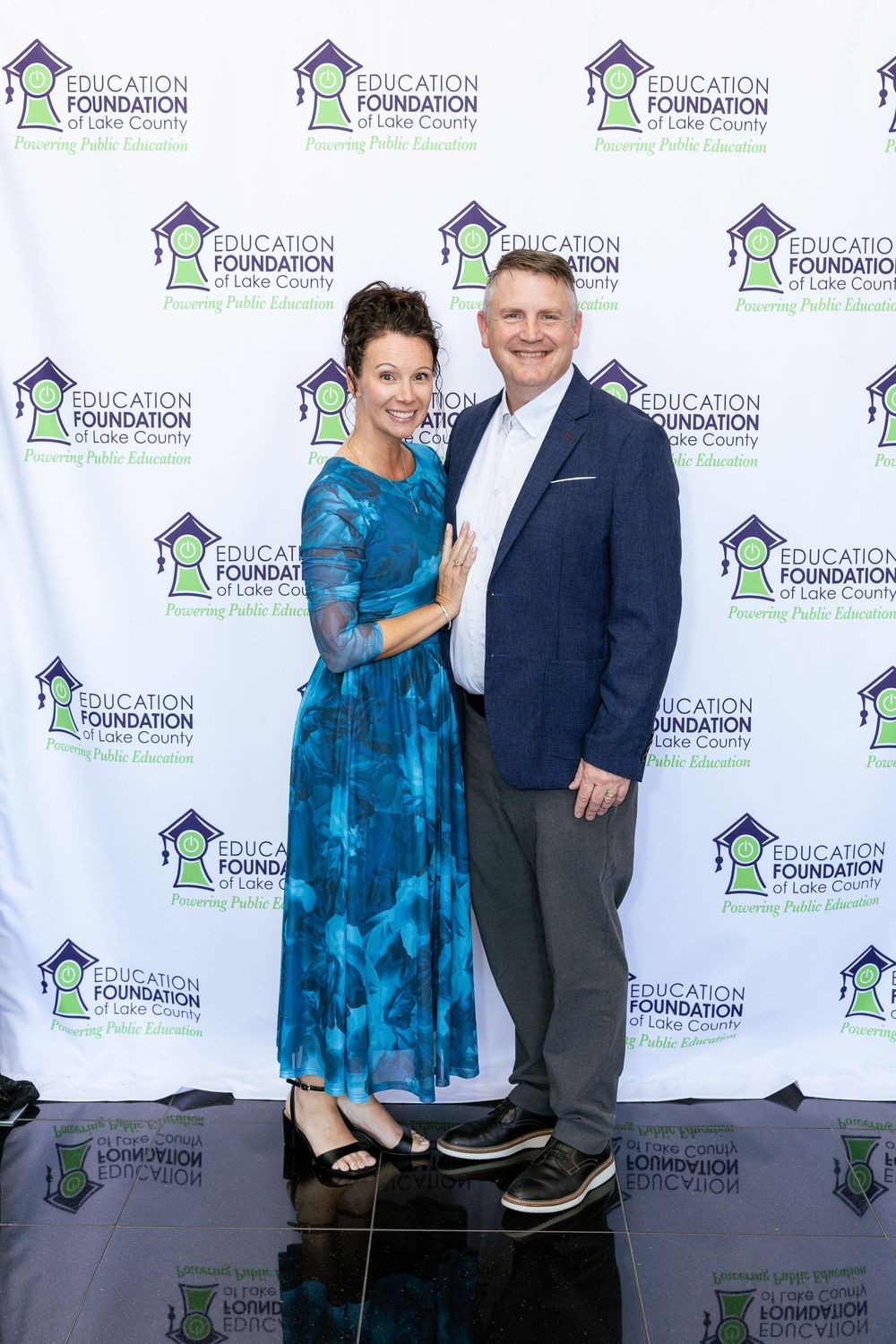 Couple posing at Education Foundation of Lake County event backdrop, woman in teal dress, man in gray suit.