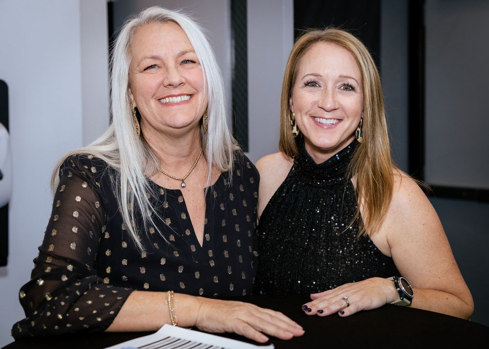 Two smiling women in black outfits pose together at a formal event, one with long white hair and one with blonde hair.