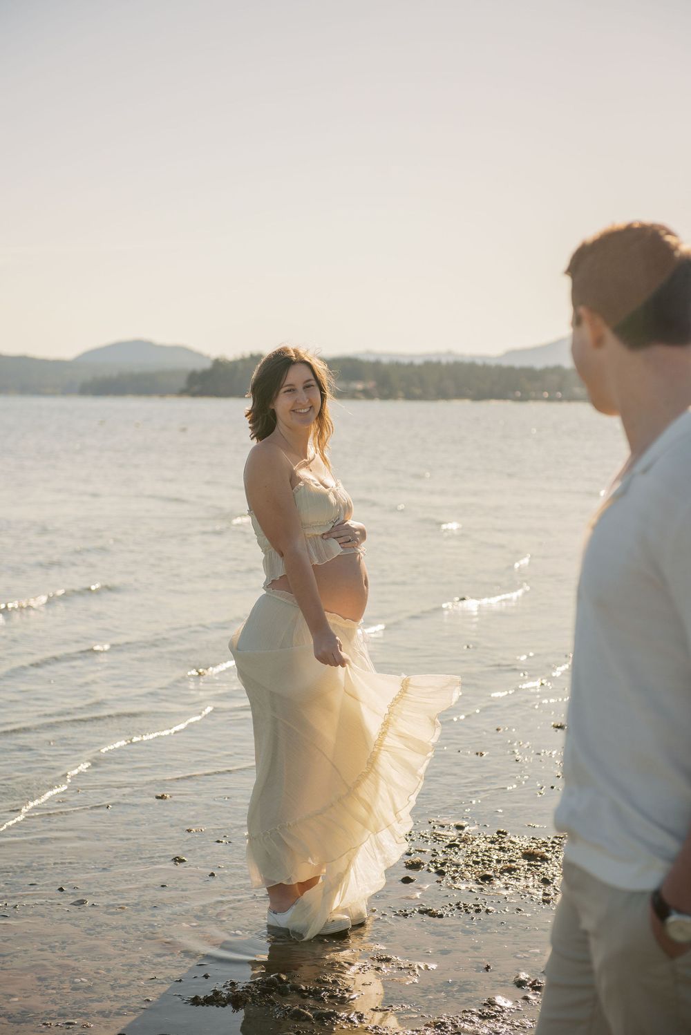 Pregnant woman in flowing cream dress standing in shallow ocean water at golden hour with partner nearby.