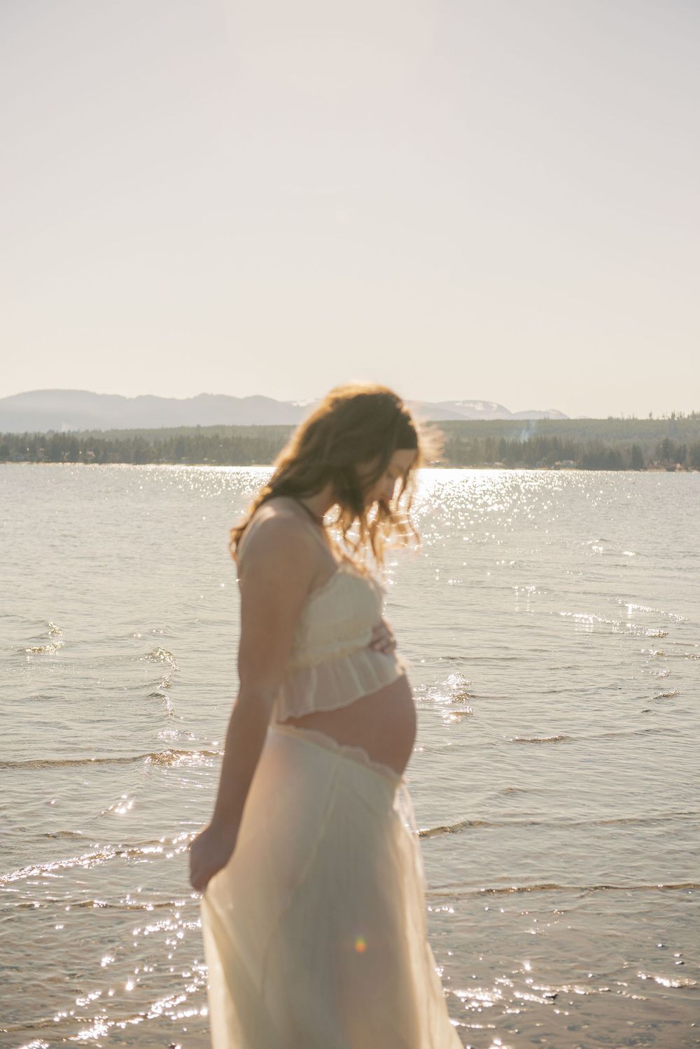 Pregnant woman in white crop top and skirt standing by a sunlit lake with mountains in the background.