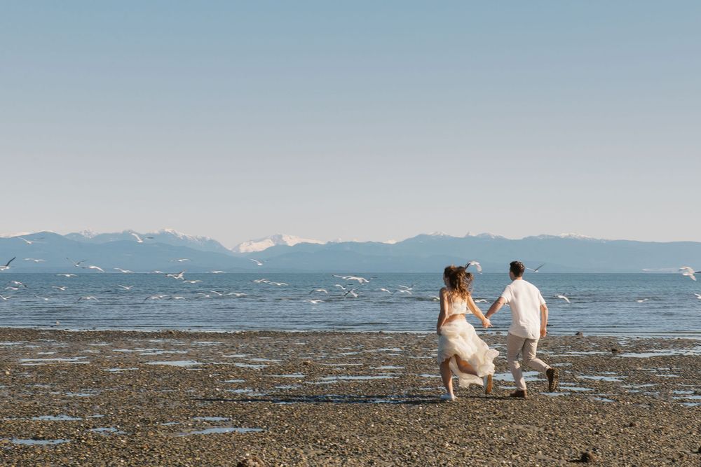 Couple in wedding attire running hand-in-hand on a rocky beach with mountain views and calm ocean waters.