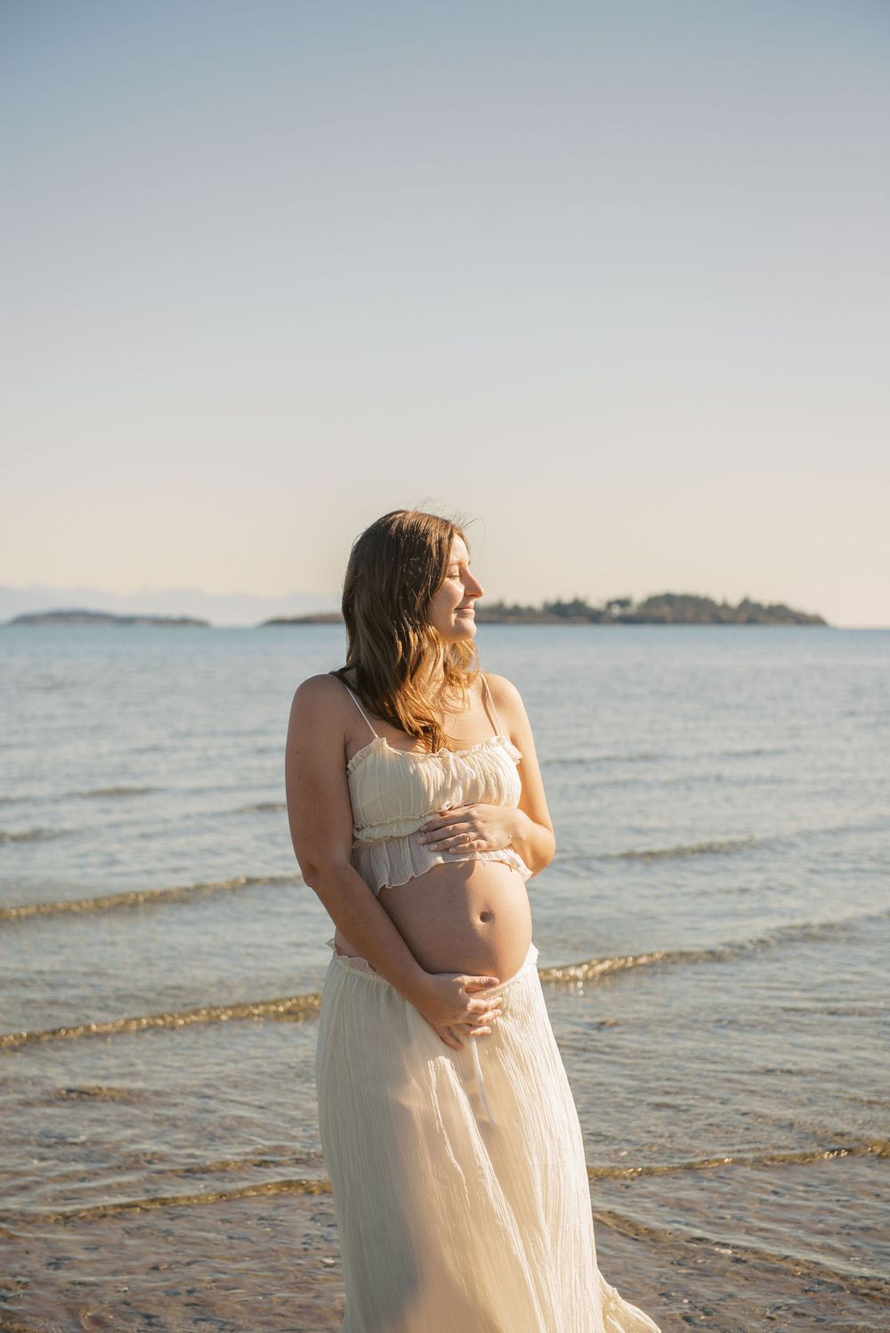Pregnant woman in white dress cradling baby bump on beach at golden hour with ocean and island in background.