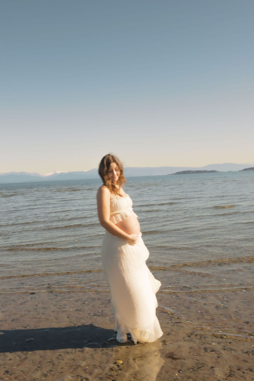 Pregnant woman in white flowing dress standing on beach shore, cradling baby bump with mountains in background.