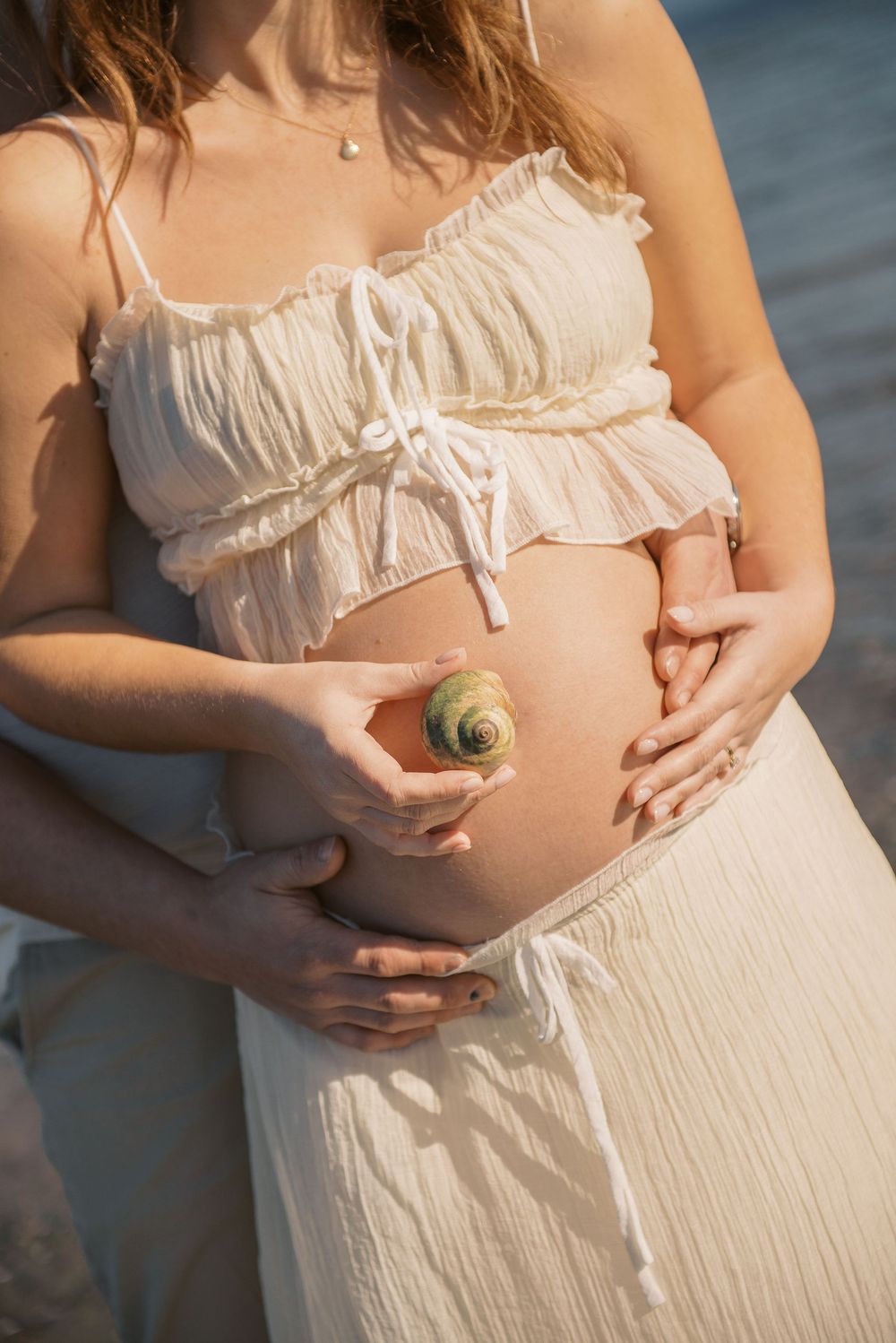 Pregnant woman in white crop top holding a seashell against her bare baby bump at the beach.