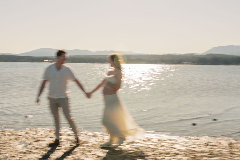 Couple holding hands walking on a sunlit beach with golden light reflecting off calm water and mountains in the background.