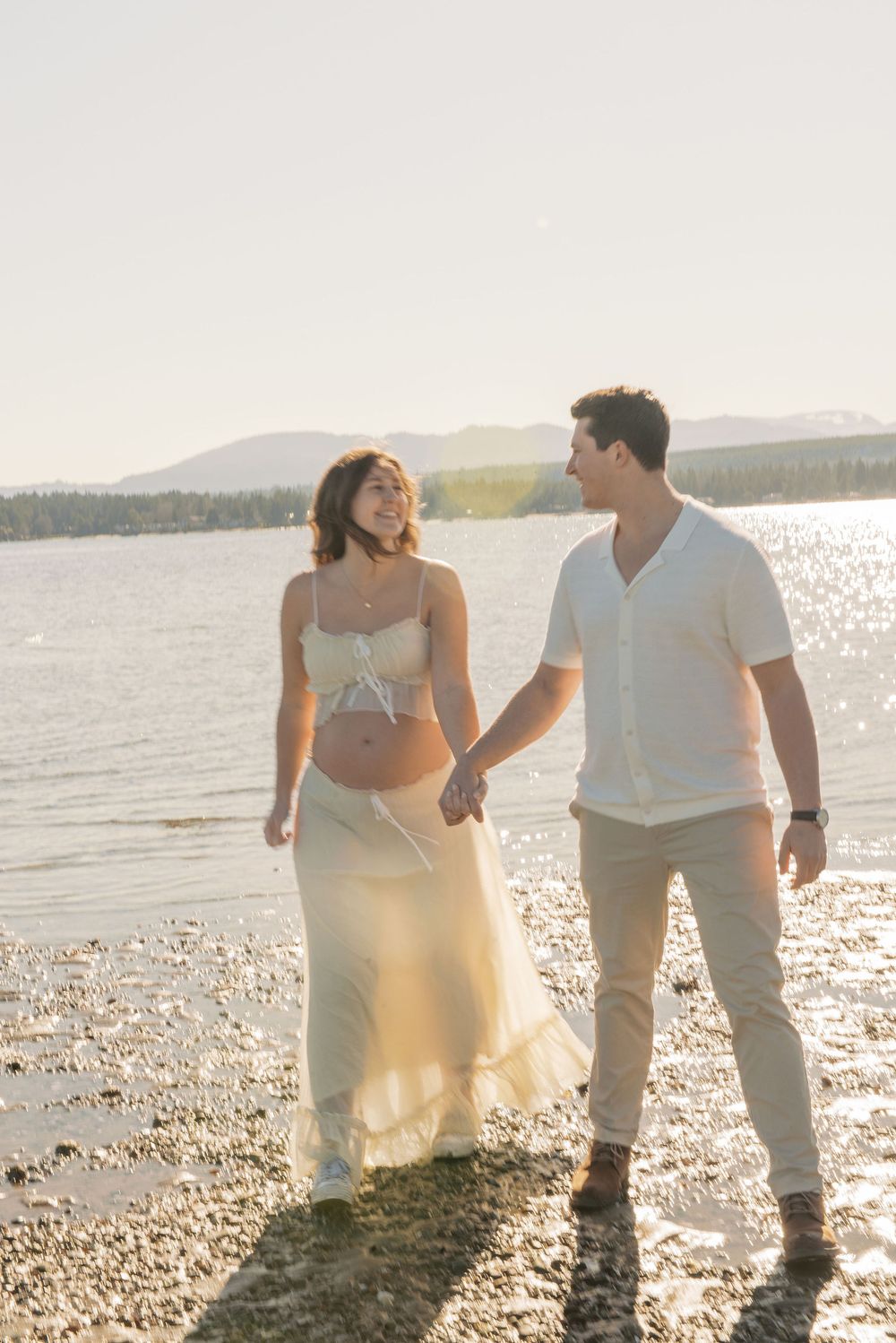 Pregnant couple holding hands and smiling on a sunny lakeside beach, woman in white crop top and skirt.