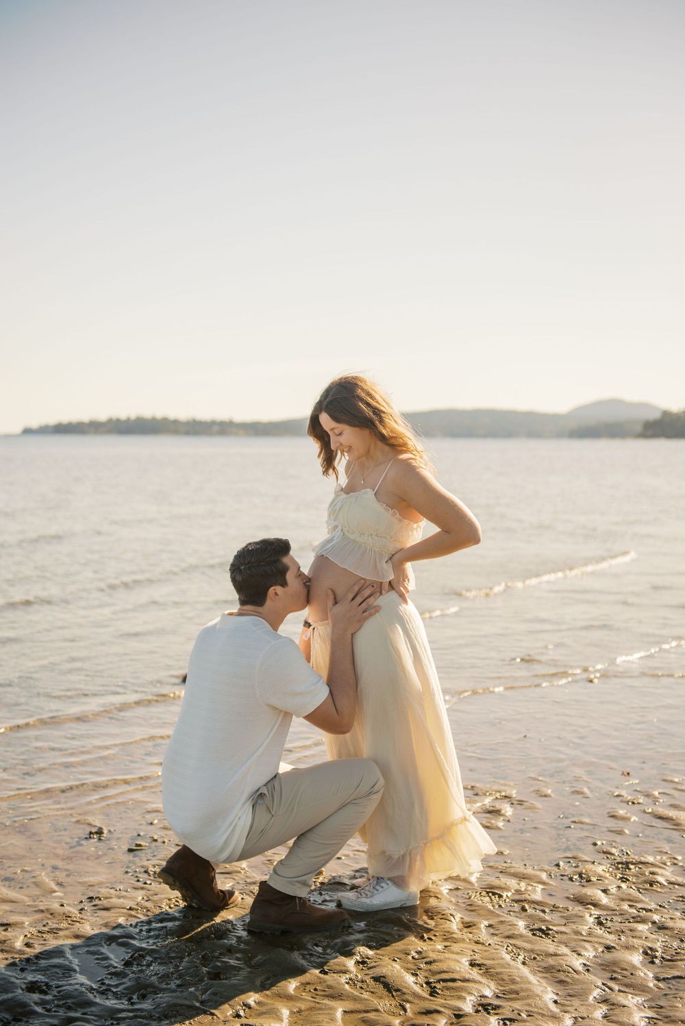 Man kneeling on sandy beach kissing pregnant woman's belly at golden hour by the ocean.
