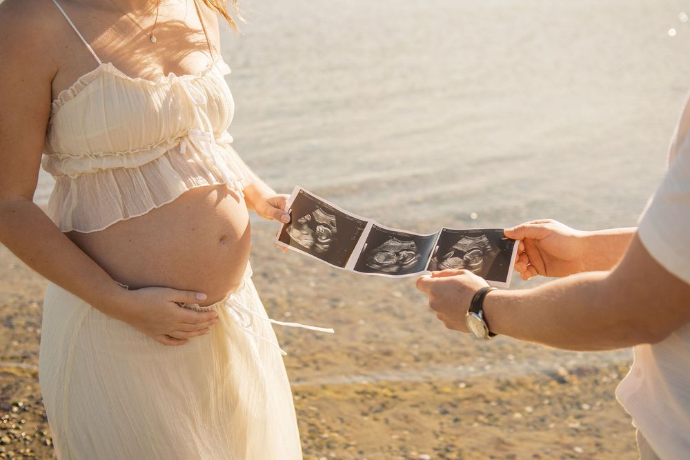 Pregnant woman in white dress holding baby ultrasound photos with partner on sunny beach.
