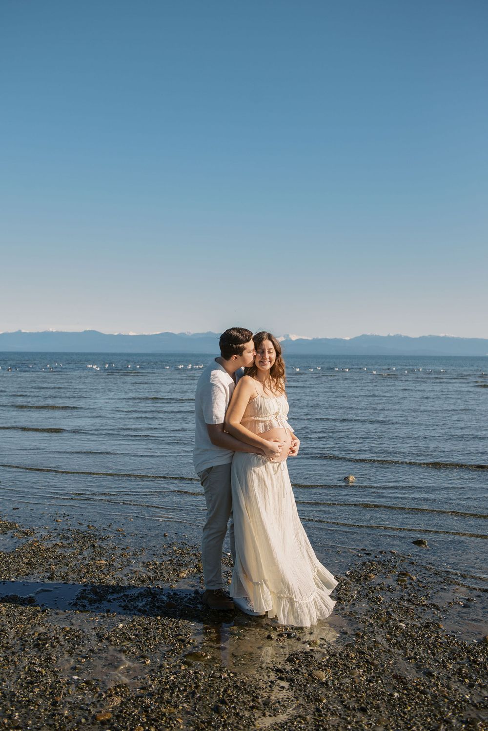 Couple embracing on rocky beach shore, pregnant woman in white dress, calm ocean and mountains in background.