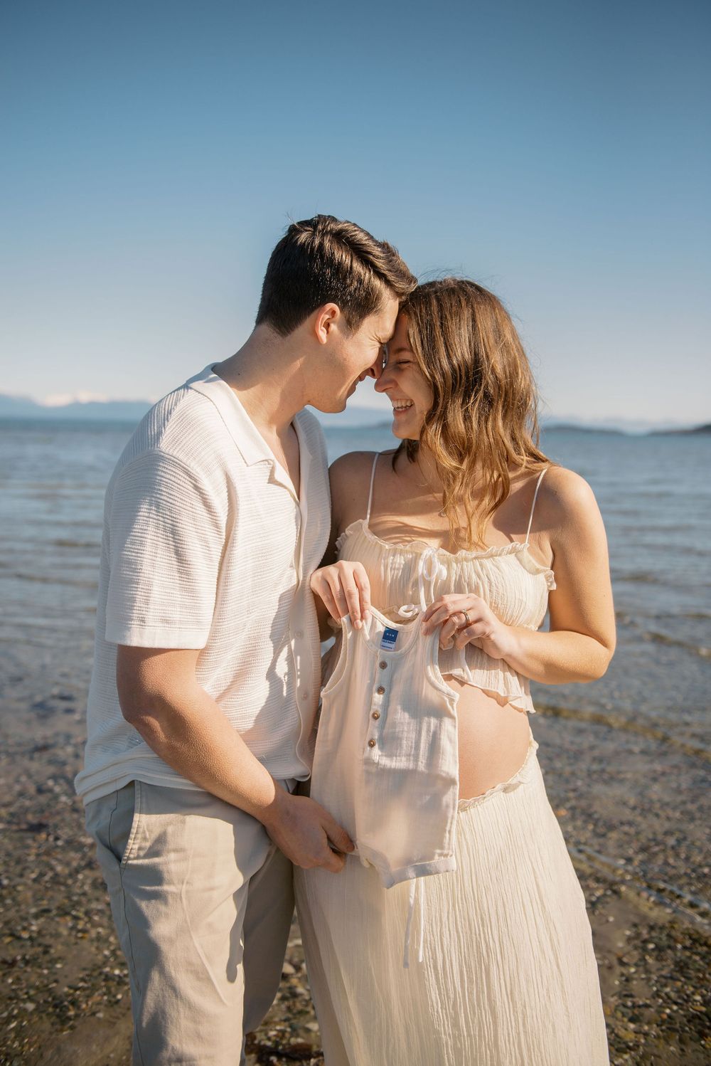 Pregnant couple standing by the lake shore holding tiny baby clothes, touching foreheads and smiling on a sunny day.